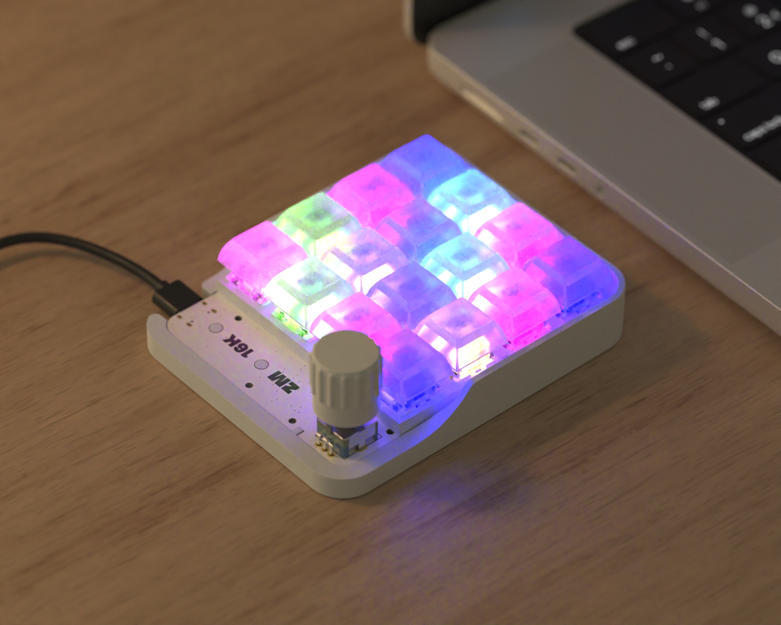 ZM16K V2 White Ghost macropad with glowing pink, purple, cyan, and green keycaps on a wooden desk beside a laptop